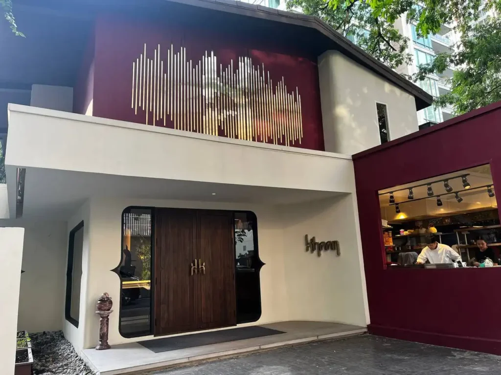 バンコクのKHAANレストラン外観。クリーム色とバーガンディのモダンな外壁に、入口上部には金色のライン装飾。右側の窓からはキッチンの様子が見える。